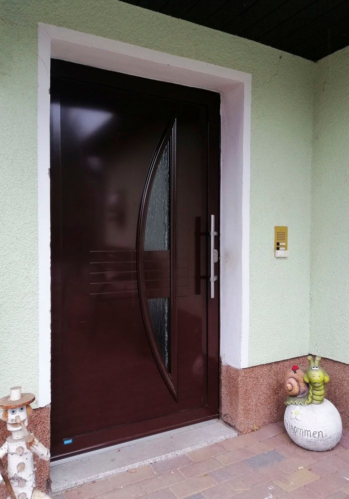 Dunkelbraune Eingangstür mit gebogener Glasscheibe neben einem an der Wand montierten Tastenfeld und Topfpflanzen am Eingang