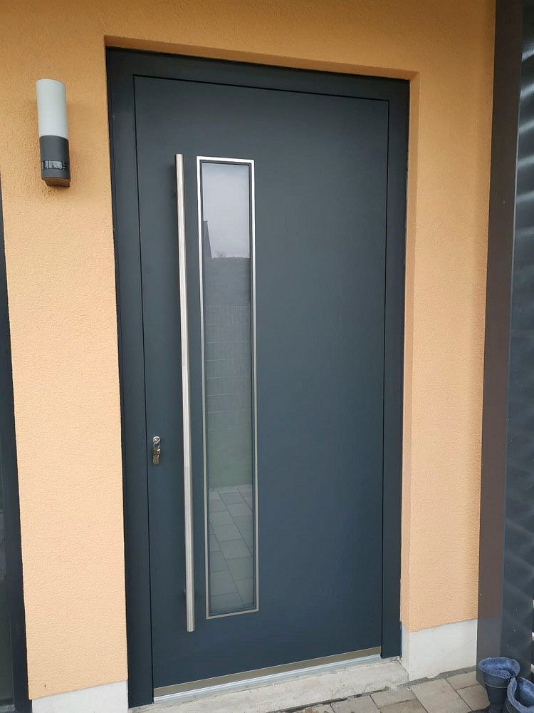 Dunkelgraue Eingangstür mit schmalem, vertikalem Fenster in einer pfirsichfarbenen Außenwand