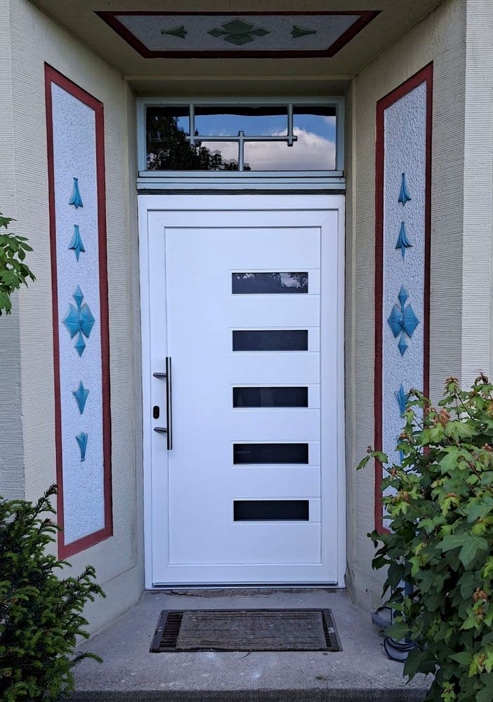 Moderne weiße Eingangstür mit horizontalen Fenstern, umrahmt von dekorativen roten und blauen Zierelementen und Grünpflanzen
