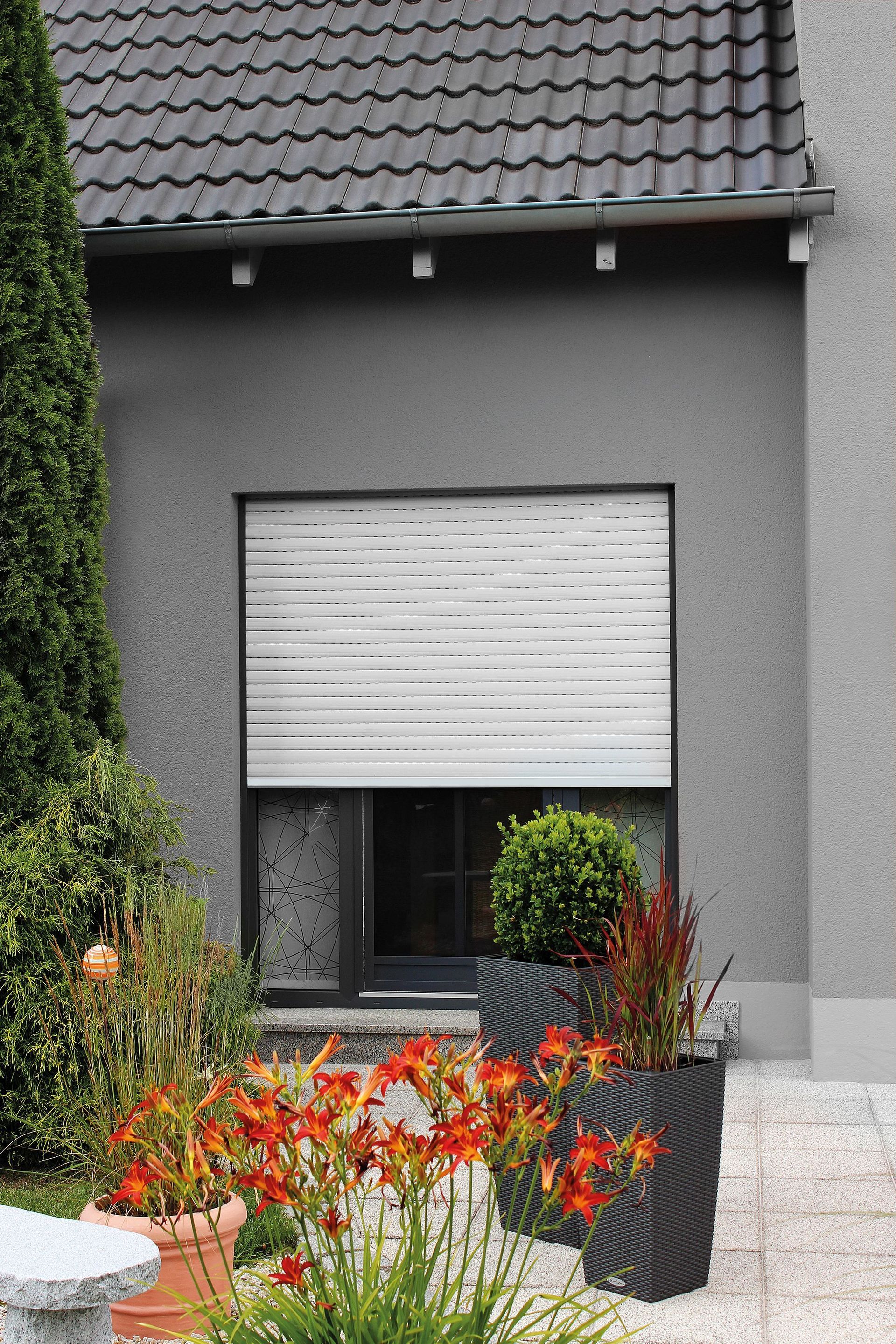 Fenster mit geschlossenen weißen Jalousien, umgeben von einem Garten mit Blumen und Topfpflanzen, an einem grauen Gebäude.