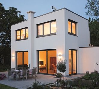 Modernes, weißes zweistöckiges Haus mit großen Fenstern, einer Terrasse und einem Garten. Warme Innenbeleuchtung bildet einen Kontrast zum Abendhimmel.