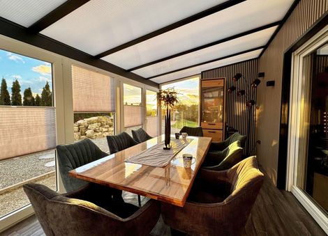 Esszimmer in einem Wintergarten mit großem Tisch, Samtstühlen und Blick auf Bäume und den Sonnenuntergang.