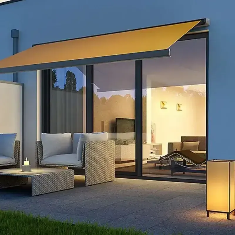 Eine Terrasse mit gelber Markise, Korbmöbeln und einer brennenden Lampe; das Innere eines Hauses ist durch Glastüren sichtbar.
