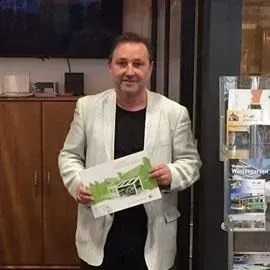 Ein Mann in einem hellgrauen Blazer hält drinnen neben einem Ausstellungsständer eine Broschüre in der Hand.