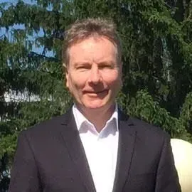 Ein lächelnder Mann in einem dunklen Anzug steht im Sonnenlicht vor Bäumen.