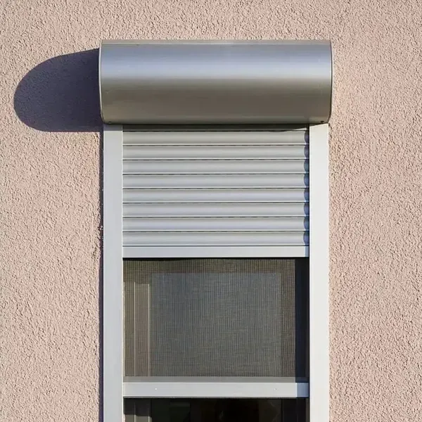 Außenfenster mit teilweise heruntergelassenem silbernen Rollladen und Fliegengitter vor einer rosa Stuckwand.