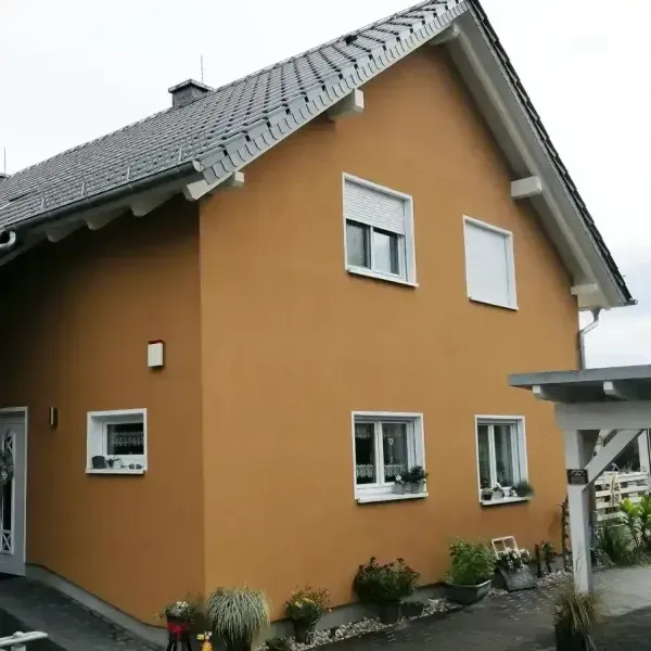 Hellbraunes zweistöckiges Haus mit grauem Dach, weißen Zierleisten und grünen Pflanzen davor.
