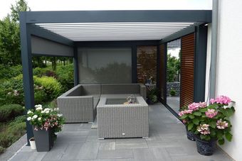 Moderne Terrasse mit grauen Korbmöbeln, rosa Topfblumen und einer dunkel umrahmten Pergola in Gartenlage