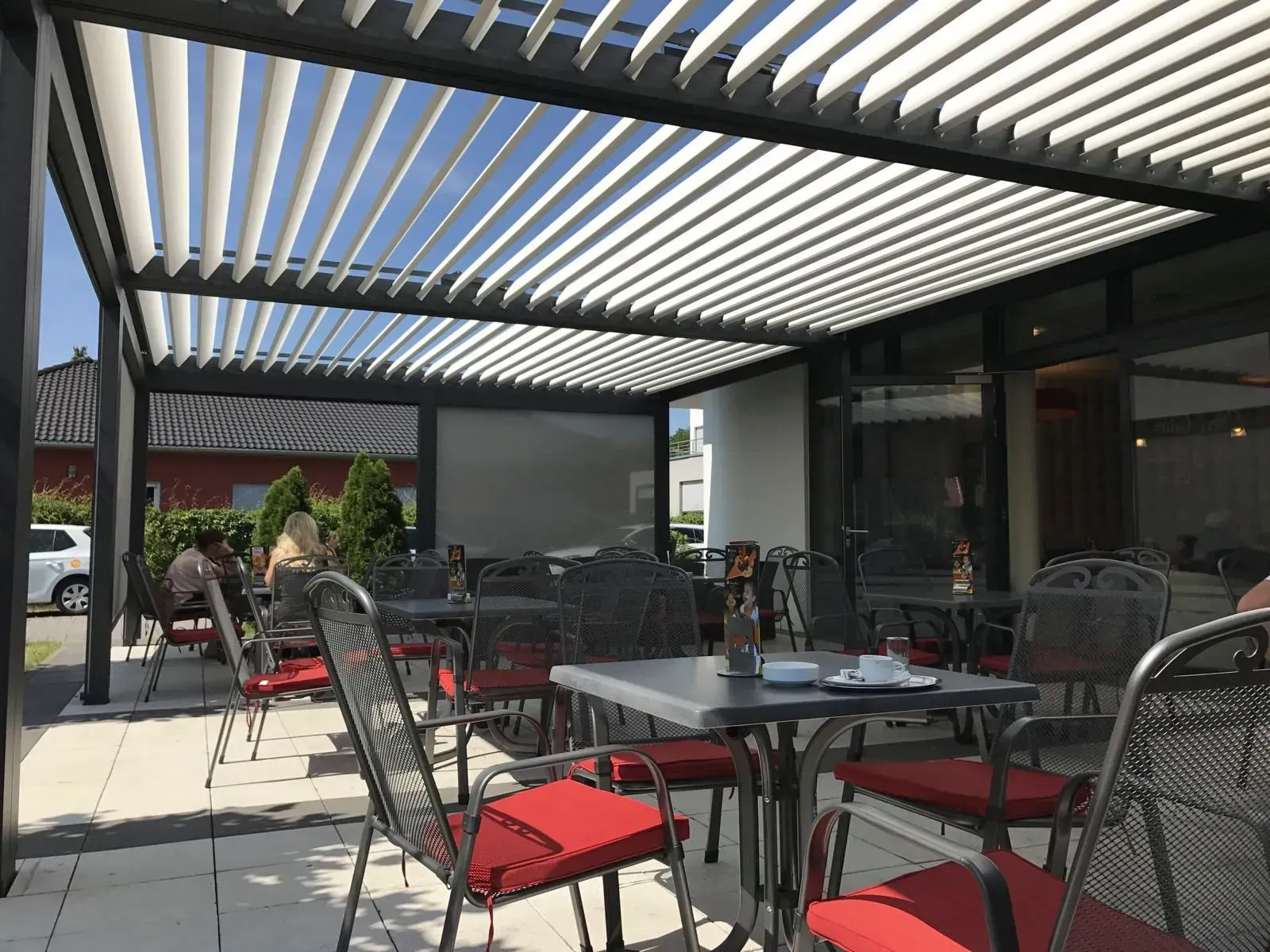 Terrasse eines Cafés im Freien mit roten Stühlen und Tischen unter einer gestreiften Pergola im hellen Sonnenlicht