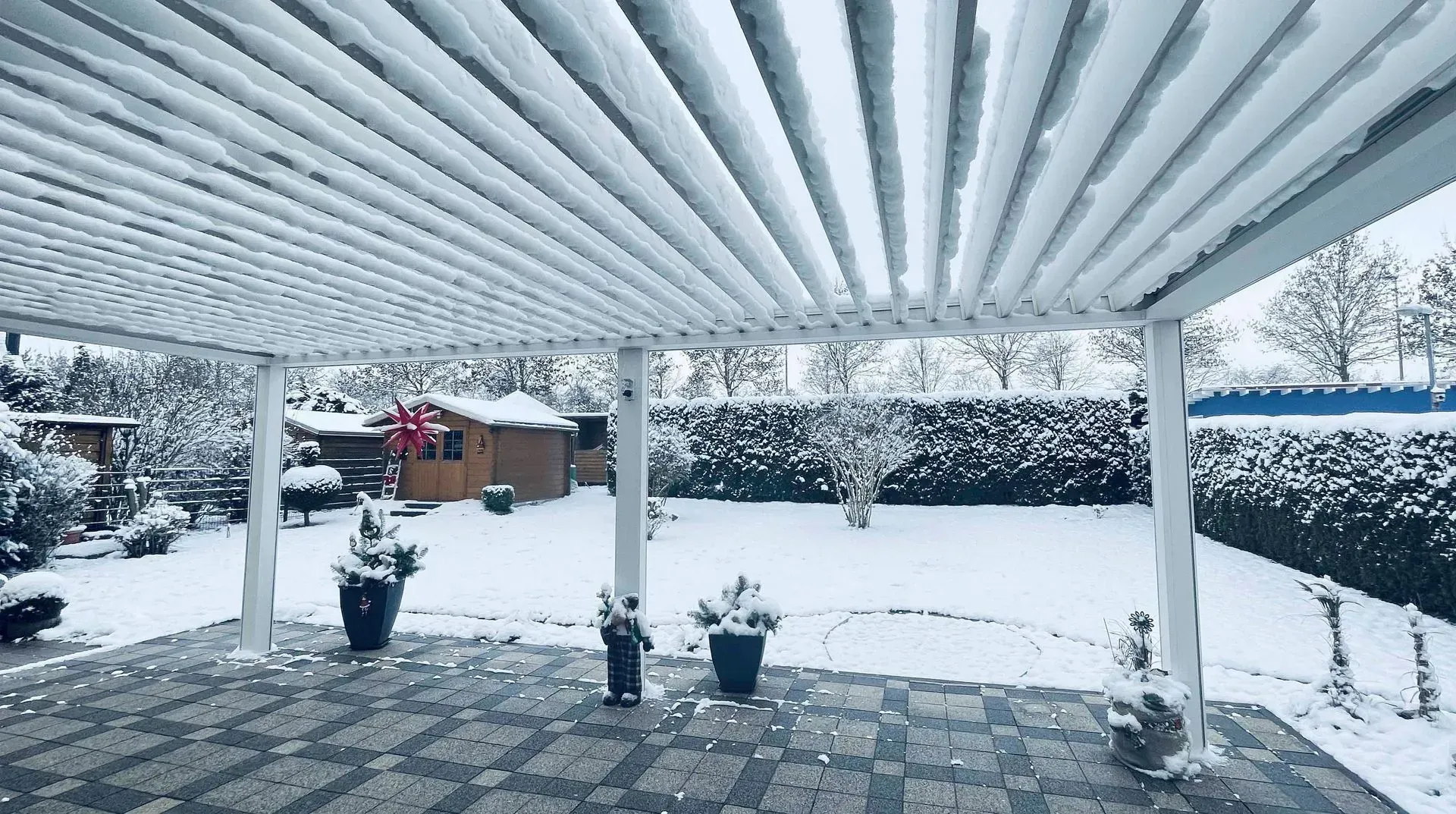 Eine schneebedeckte Terrasse mit Pergola, Topfpflanzen und ein schneebedeckter Garten.