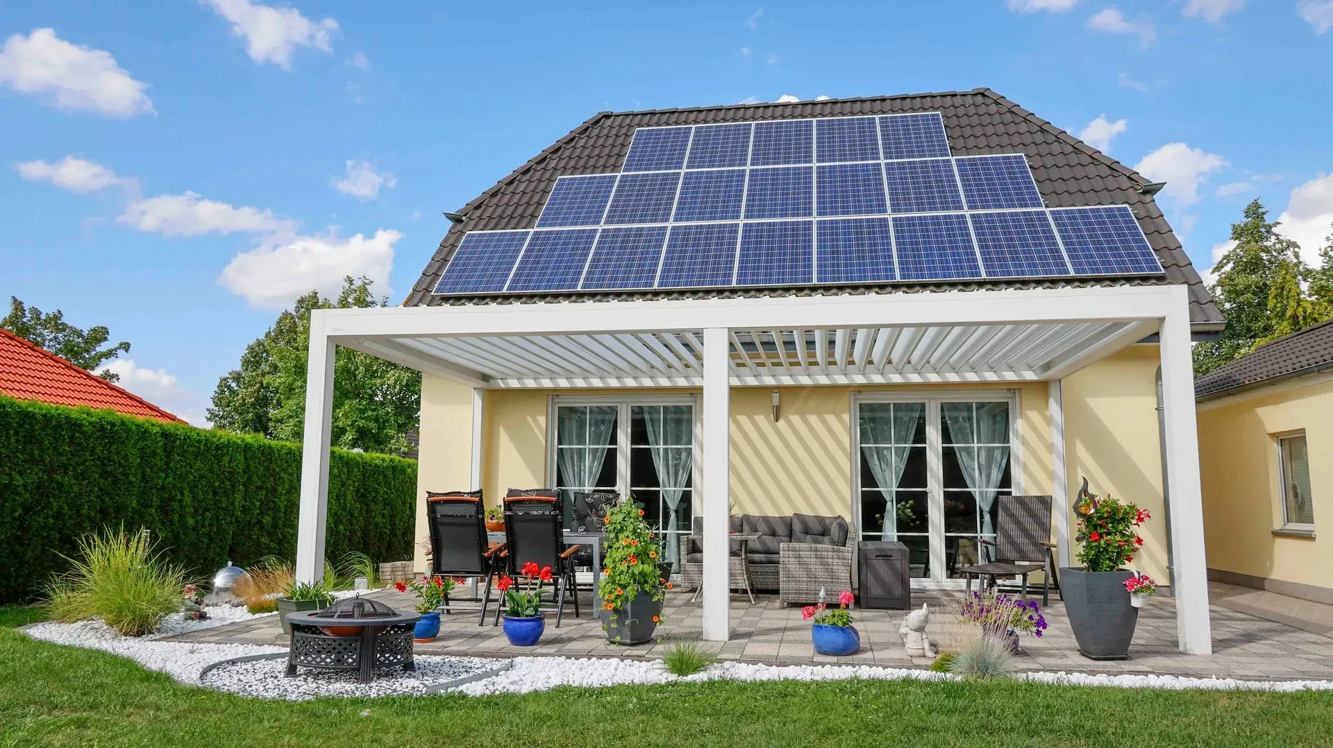 Haus mit Solarpaneelen und überdachter Terrasse mit Stühlen und Topfpflanzen unter blauem Himmel