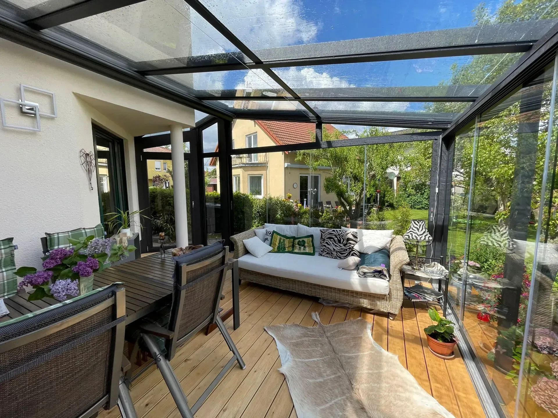 Überdachte Terrasse mit Korbmöbeln, Esstisch und Topfpflanzen neben einem Haus, darüber ein Glasdach.