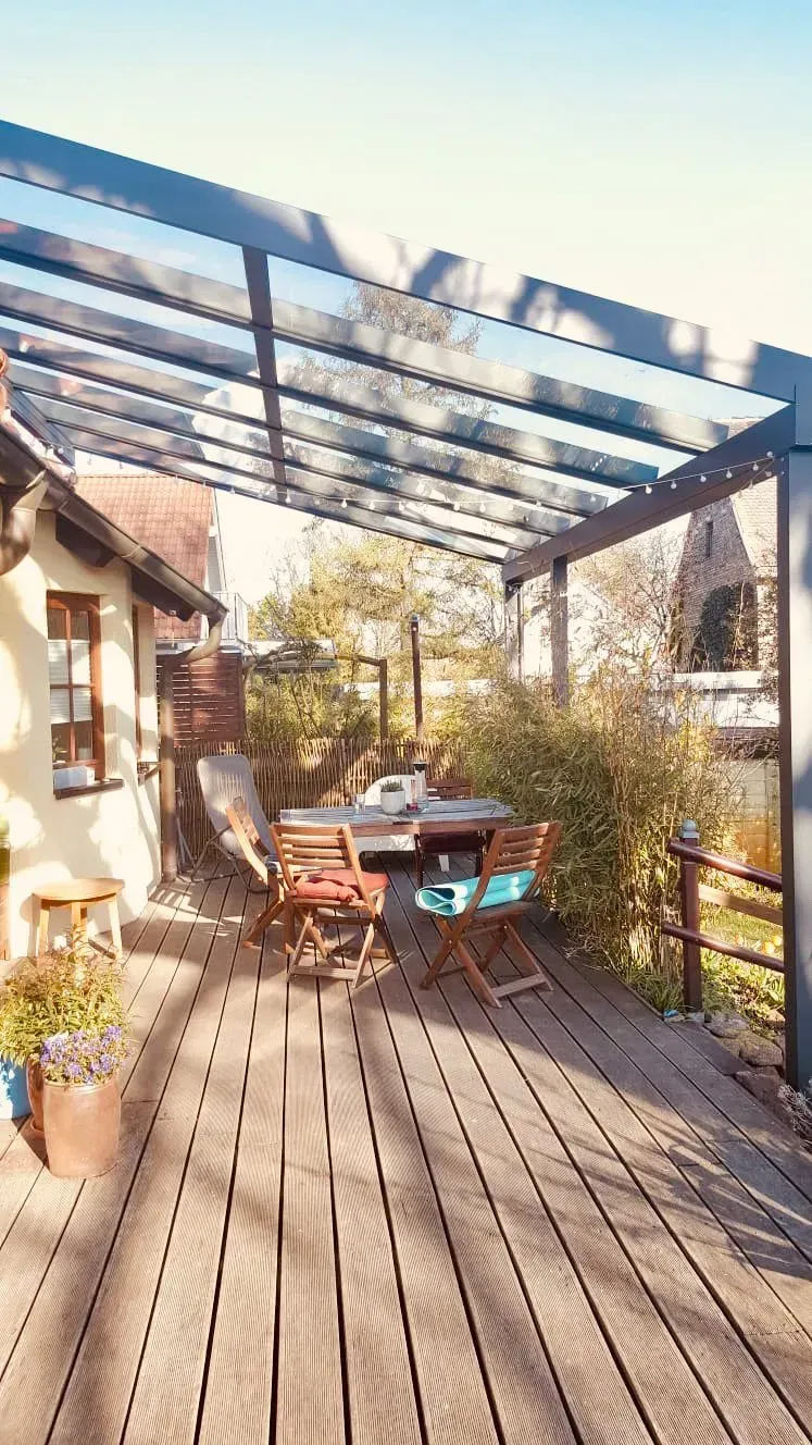 Sonnige Holzterrasse mit Terrassentisch und Stühlen unter einer Glaspergola neben einem Haus