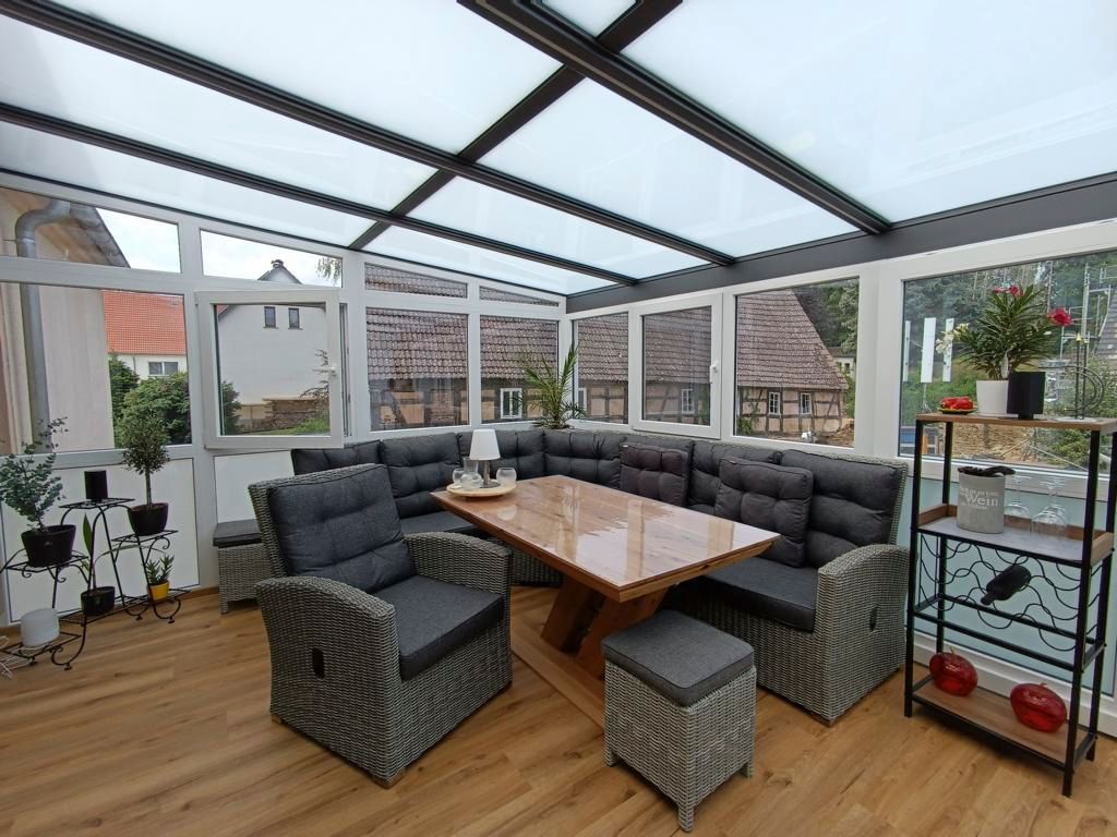 Heller Wintergarten mit grauer Eckcouch, Holztisch, Korbsessel und Holzboden unter einem Glasdach