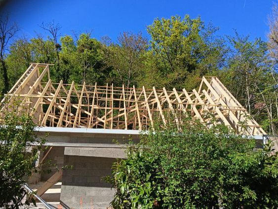 Une charpente de toit en bois en construction sur une base en béton, se détachant sur un fond d'arbres.