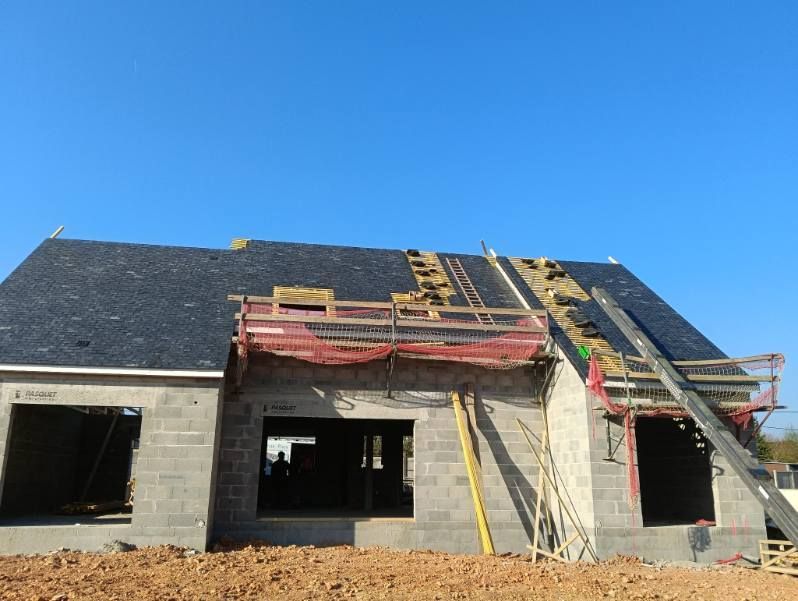 Un chantier de construction montrant une maison aux murs en béton, un toit sombre partiellement recouvert de tuiles et des échafaudages.
