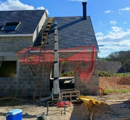 Une maison en pierre neuve en construction, avec un toit en bardeaux sombres, une cheminée et un filet de sécurité au-dessus de l'entrée.