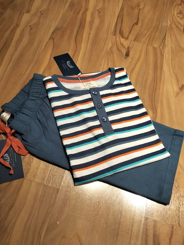 Una camiseta tipo Henley a rayas naranja, azul y blanca, doblada, descansa sobre unos pantalones azules de estar por casa en un suelo de madera.