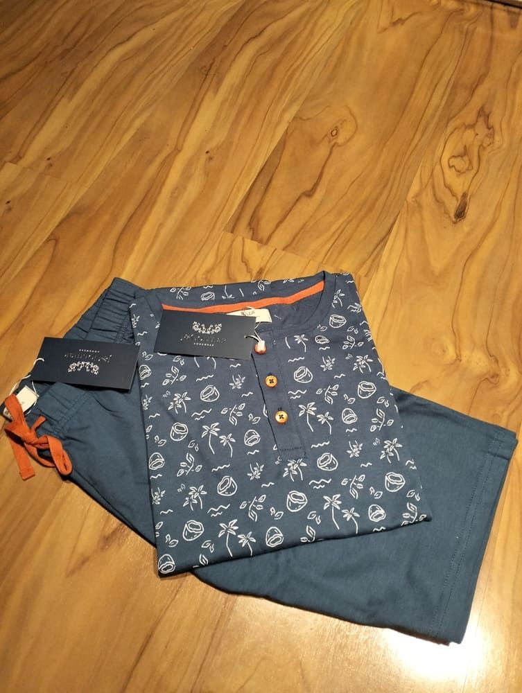 Una camisa Henley azul estampada, doblada y con cordones naranjas, descansa sobre unos pantalones cortos a juego, sobre un fondo de madera.