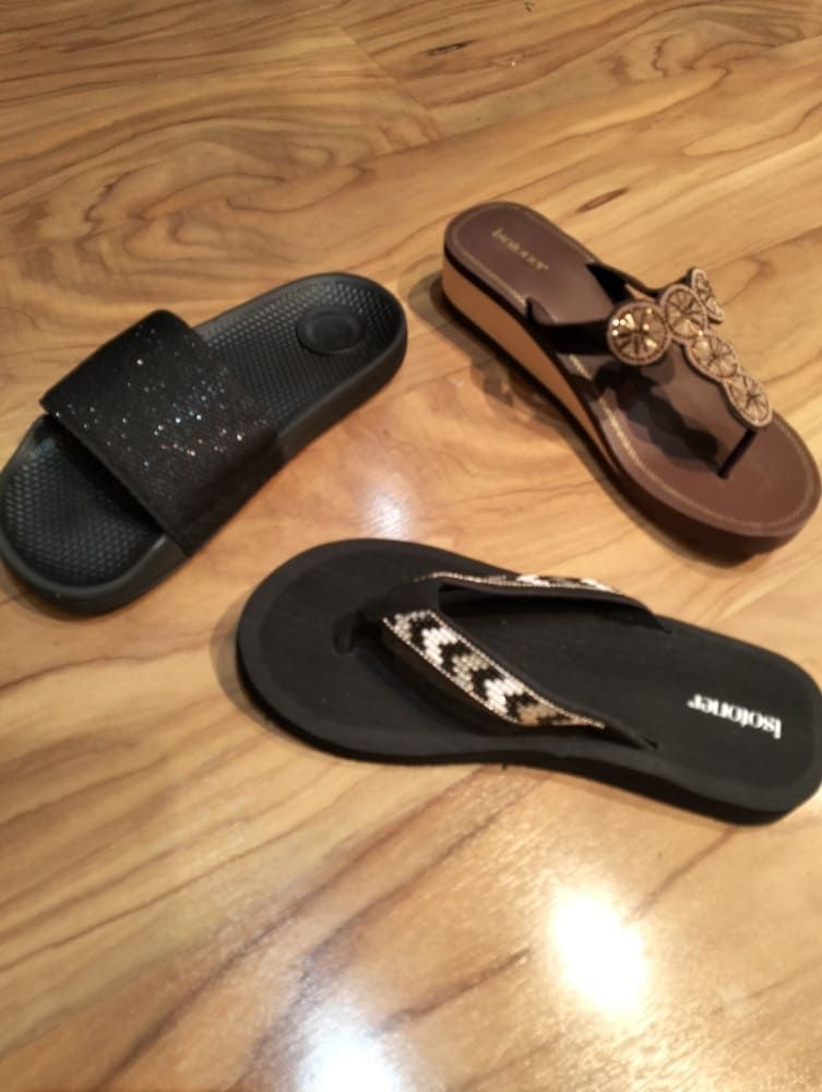 Tres pares de sandalias, entre ellas una sandalia negra tipo pala, una cuña marrón adornada y una chancla negra, sobre un suelo de madera.