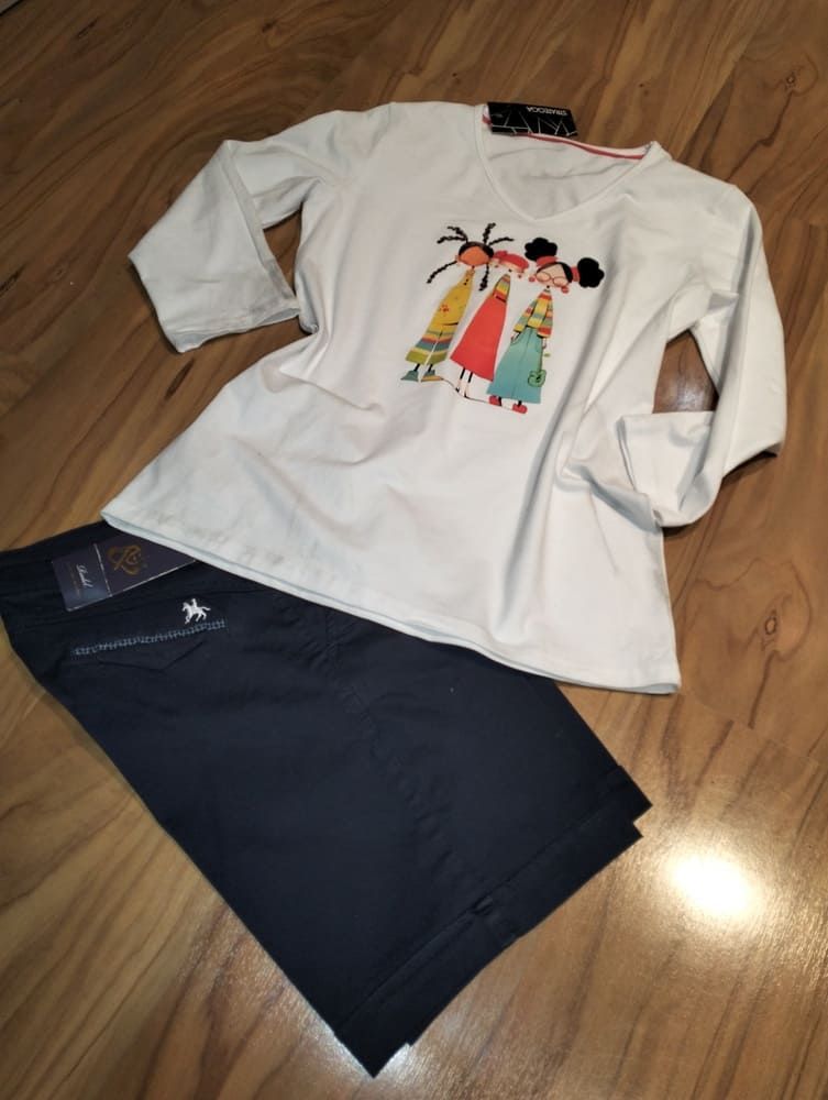 Una camisa blanca de manga larga con un estampado de dibujos animados de tres figuras yace junto a unos pantalones cortos azul oscuro sobre una superficie de madera.