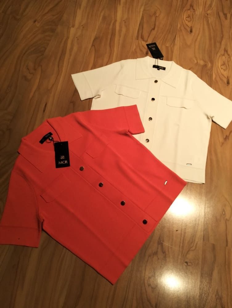 Dos camisas de punto de manga corta con botones, una de color coral y otra blanca, extendidas sobre un suelo de madera con las etiquetas de la tienda aún puestas.
