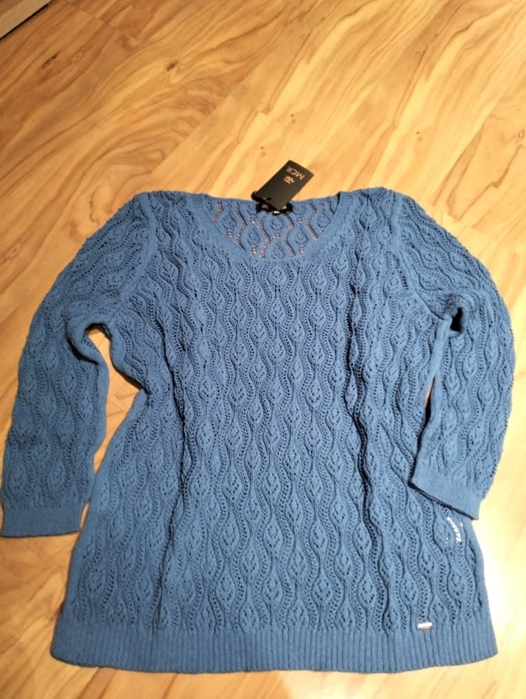 Un suéter azul de manga larga y punto calado con un estampado de rombos texturizados, extendido sobre un suelo de madera clara.