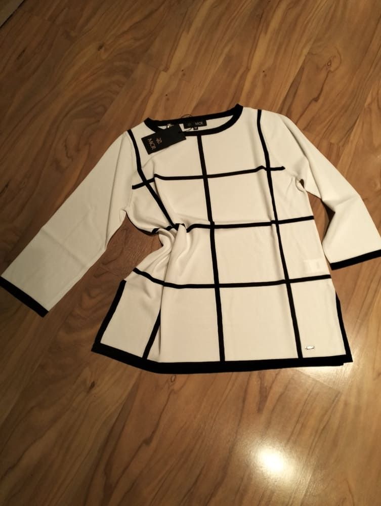 Un jersey blanco de manga larga con un estampado de cuadrícula negra, que se muestra extendido sobre un suelo de madera.