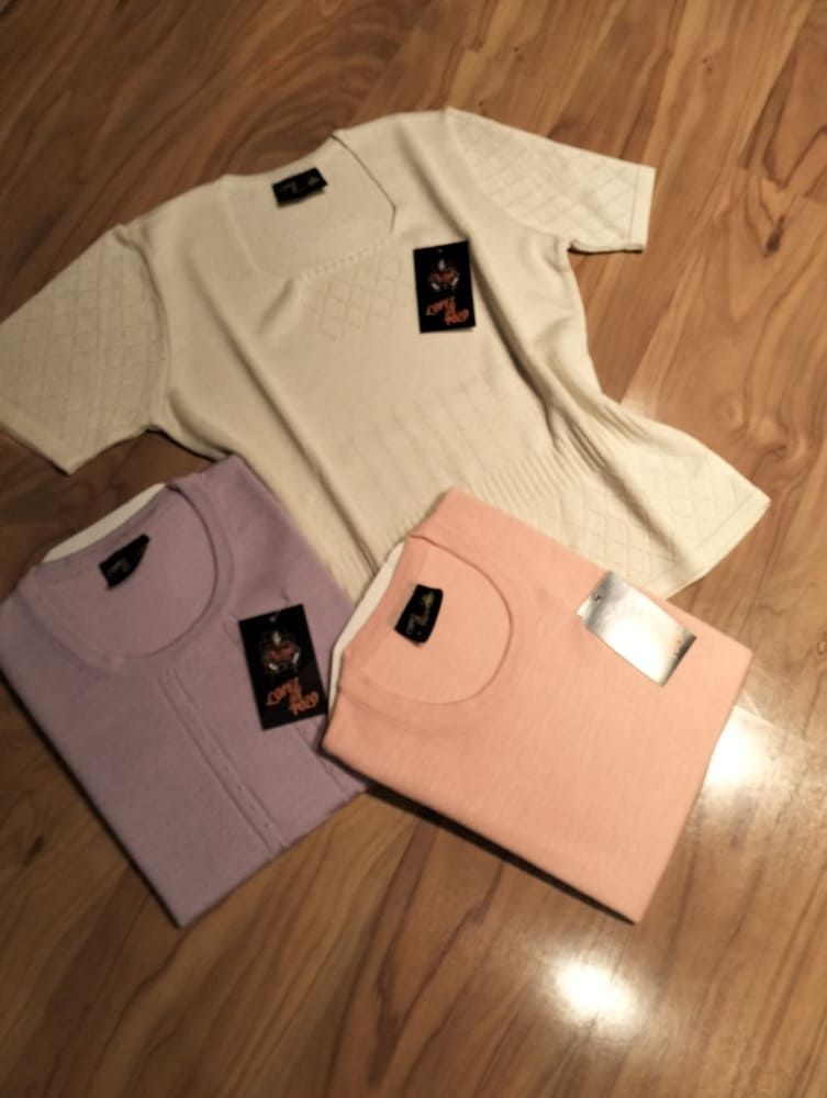 Tres camisas de manga corta y tejido acanalado, en color crema, lavanda y melocotón, cada una con una etiqueta de la marca, estaban dispuestas sobre un suelo de madera.