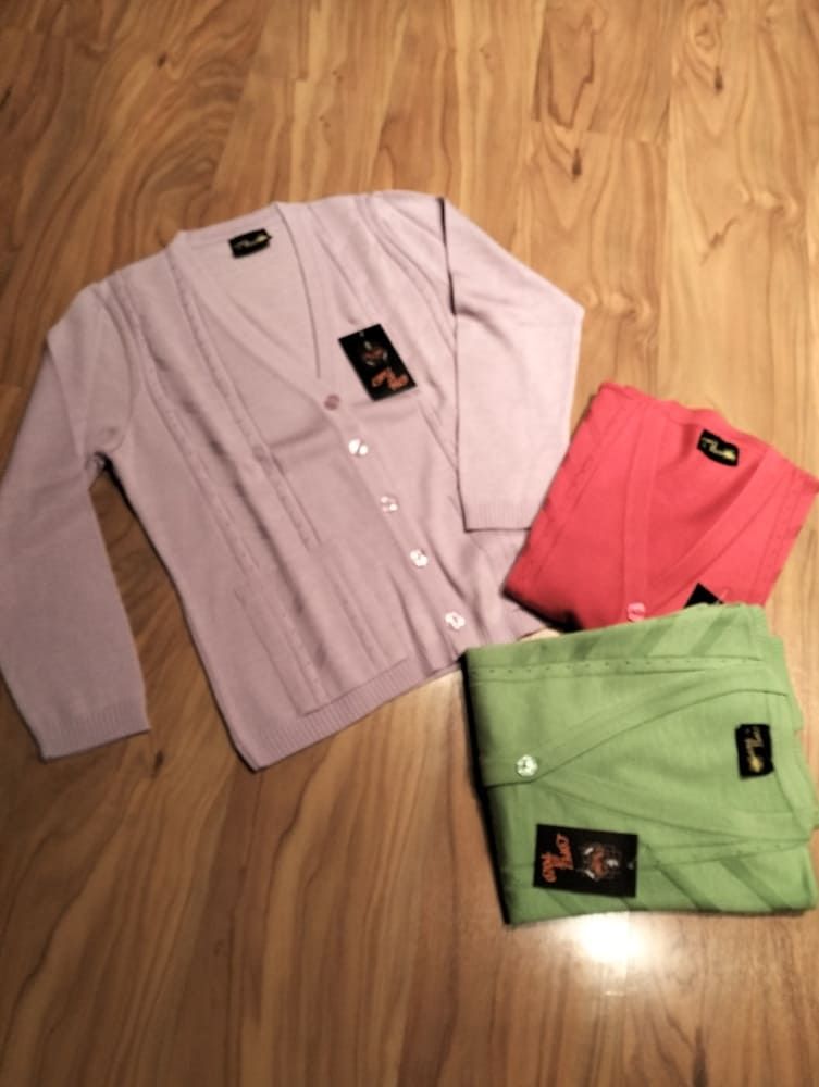 Tres cárdigans con escote en V en color lila, coral y verde claro, extendidos sobre un suelo de madera de color claro.