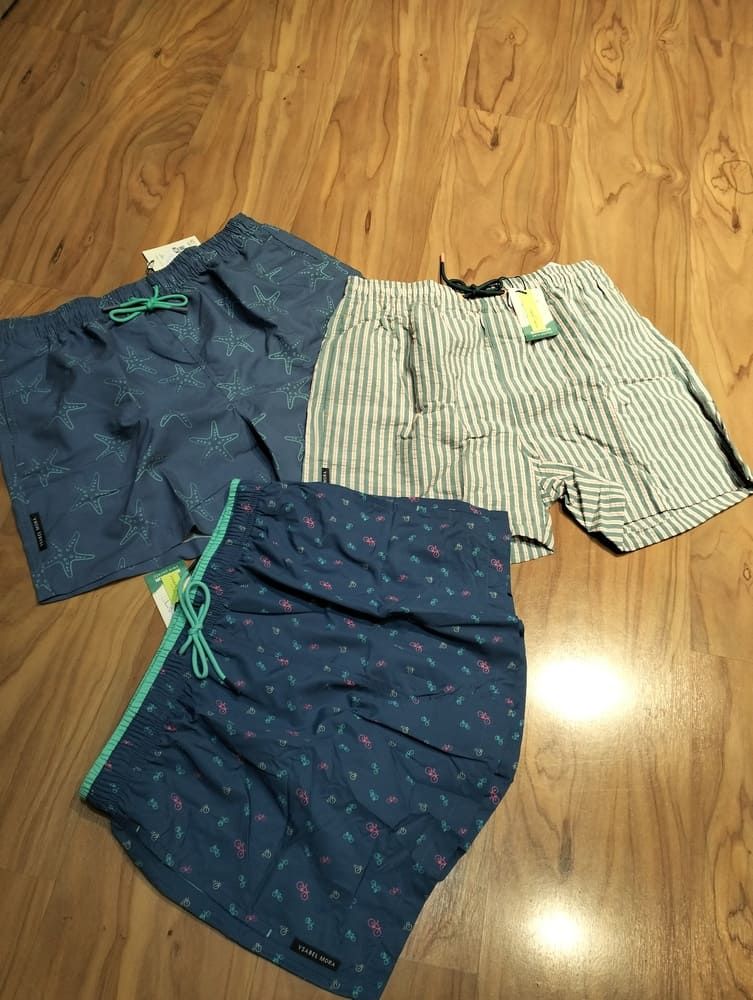 Tres pares de bañadores dispuestos sobre un suelo de madera: uno azul liso, uno a rayas y otro con una cinturilla de color verde azulado.