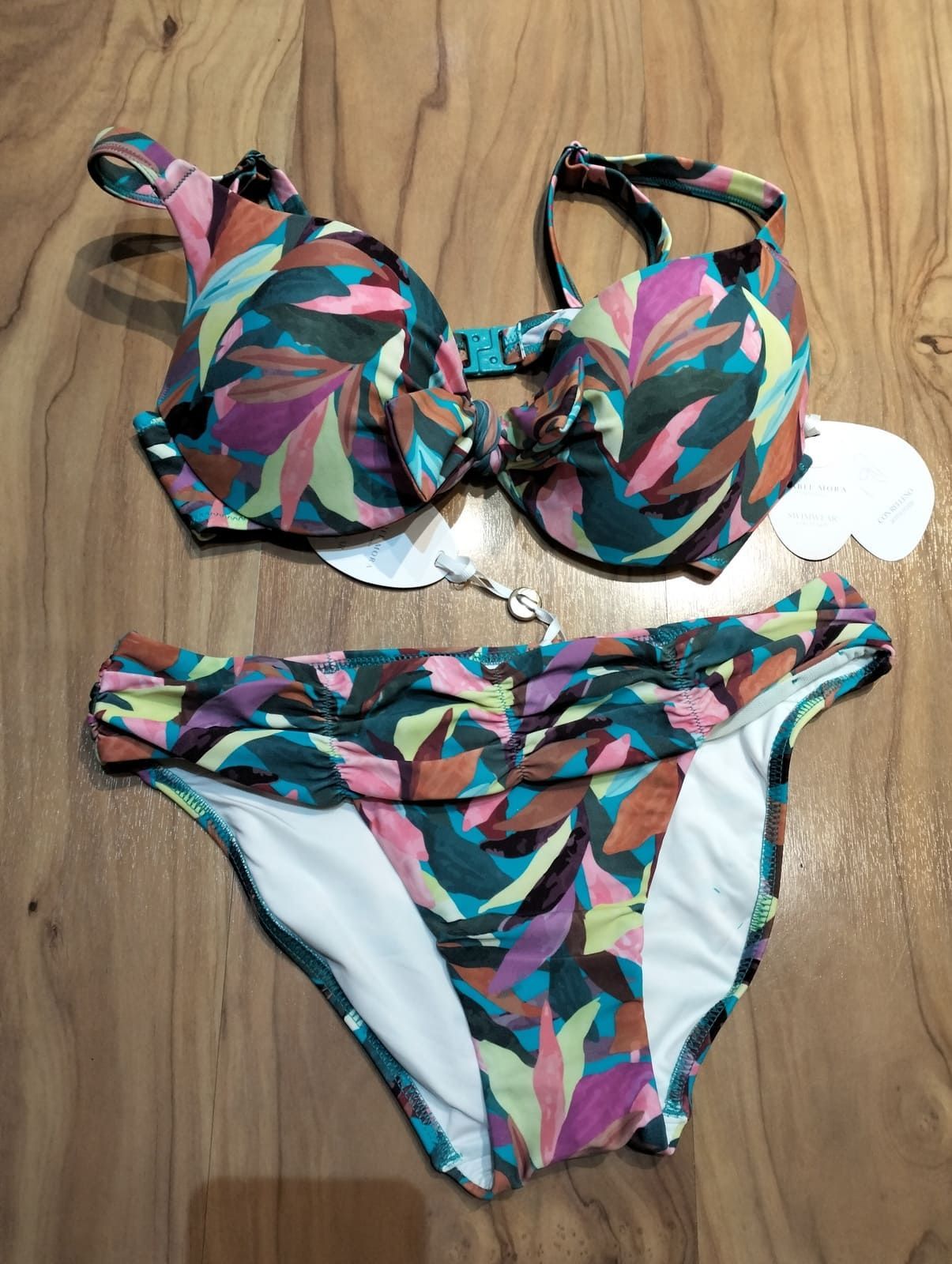 Un bikini de dos piezas con un estampado botánico de hojas oscuras en tonos verdes, morados y amarillos, extendido sobre una superficie de madera.