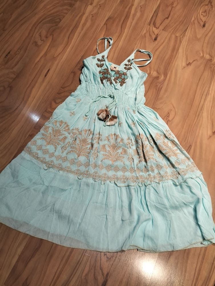 Un vestido de verano blanco, sin mangas, de largo midi, con ribete de encaje y detalles escalonados, extendido sobre un suelo de madera clara.