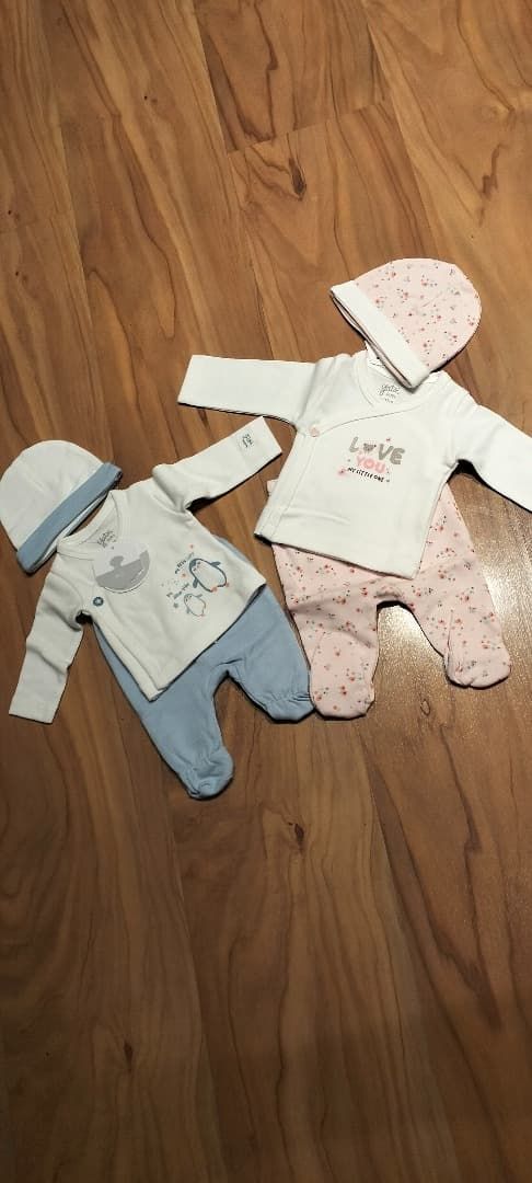 Dos conjuntos de bebé con gorritos, uno azul y otro rosa con lunares, colocados sobre un suelo de madera