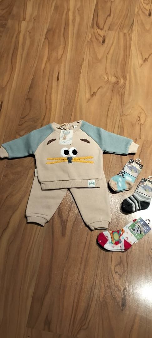 Conjunto de ropa para bebé con camiseta color crema y azul con estampado de animales, pantalón a rayas, gorro a juego y calcetines sobre un suelo de madera