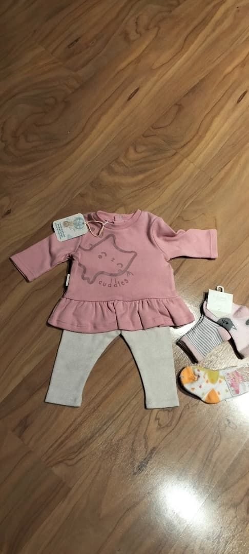 Vestido rosa para bebé con leggings grises y un pequeño conjunto de bebé sobre un suelo de madera