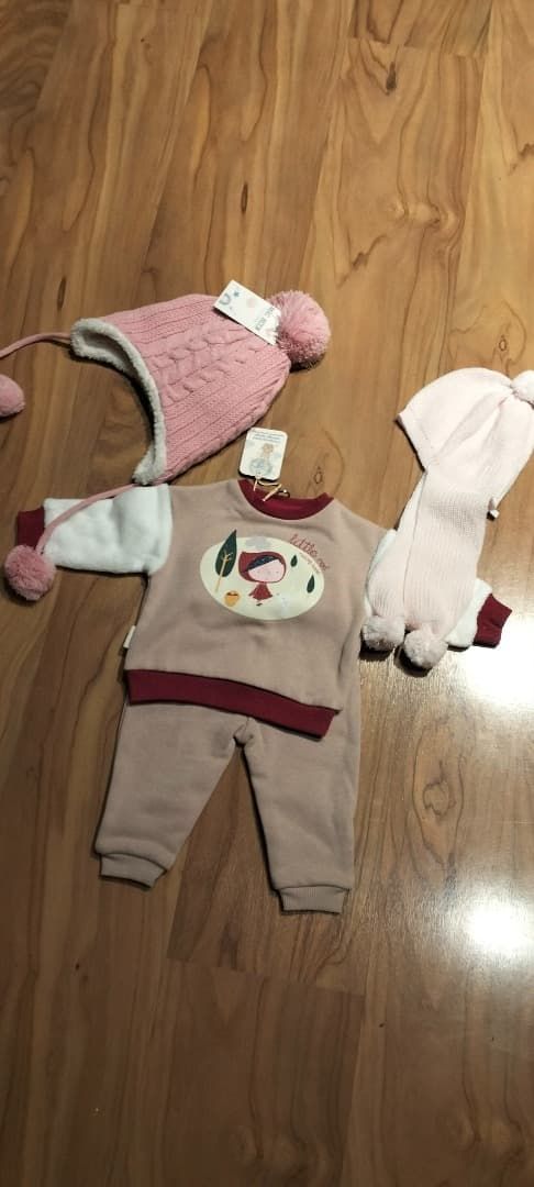 Tres conjuntos de bebé colocados sobre un suelo de madera, entre ellos un conjunto de punto rosa y un pelele tipo jersey beige.