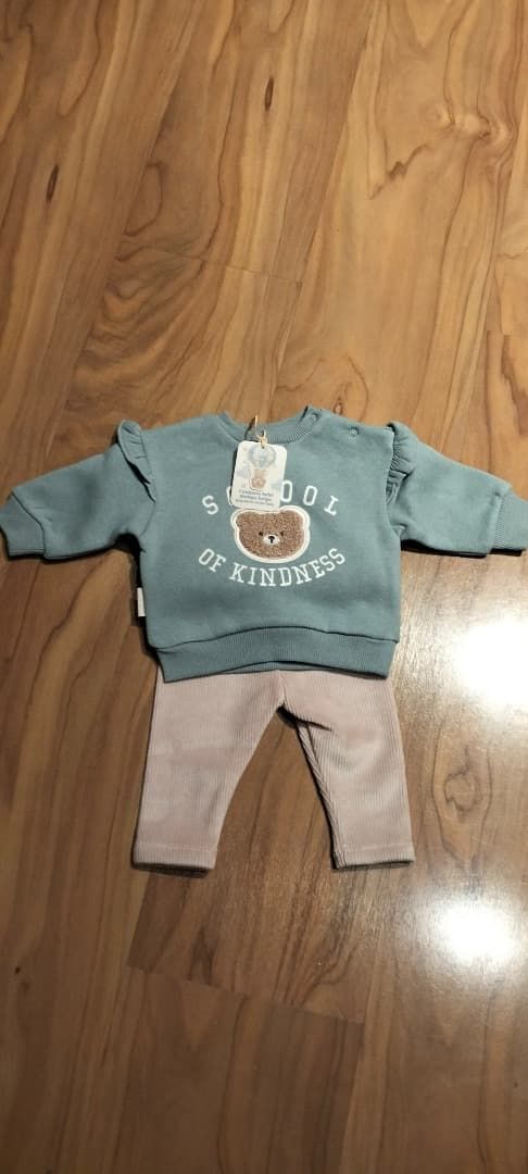 Conjunto infantil sobre un suelo de madera: sudadera verde azulado con un oso y pantalones gris claro.