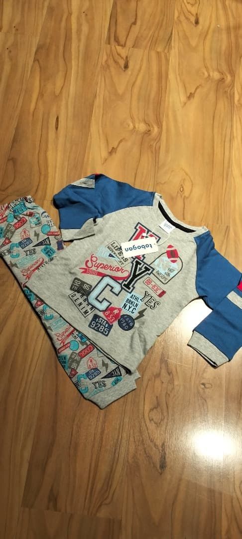 Pijama infantil extendido sobre un suelo de madera, con una camiseta gris estampada y mangas azules, y un pantalón estampado.