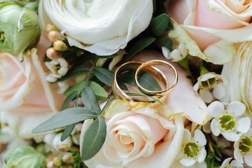 Zwei goldene Eheringe liegen auf einem Blumenarrangement aus weißen und rosafarbenen Rosen sowie grünen Blättern. Zwei goldene Eheringe liegen auf einem Blumenarrangement aus weißen und rosafarbenen Rosen sowie grünen Blättern.