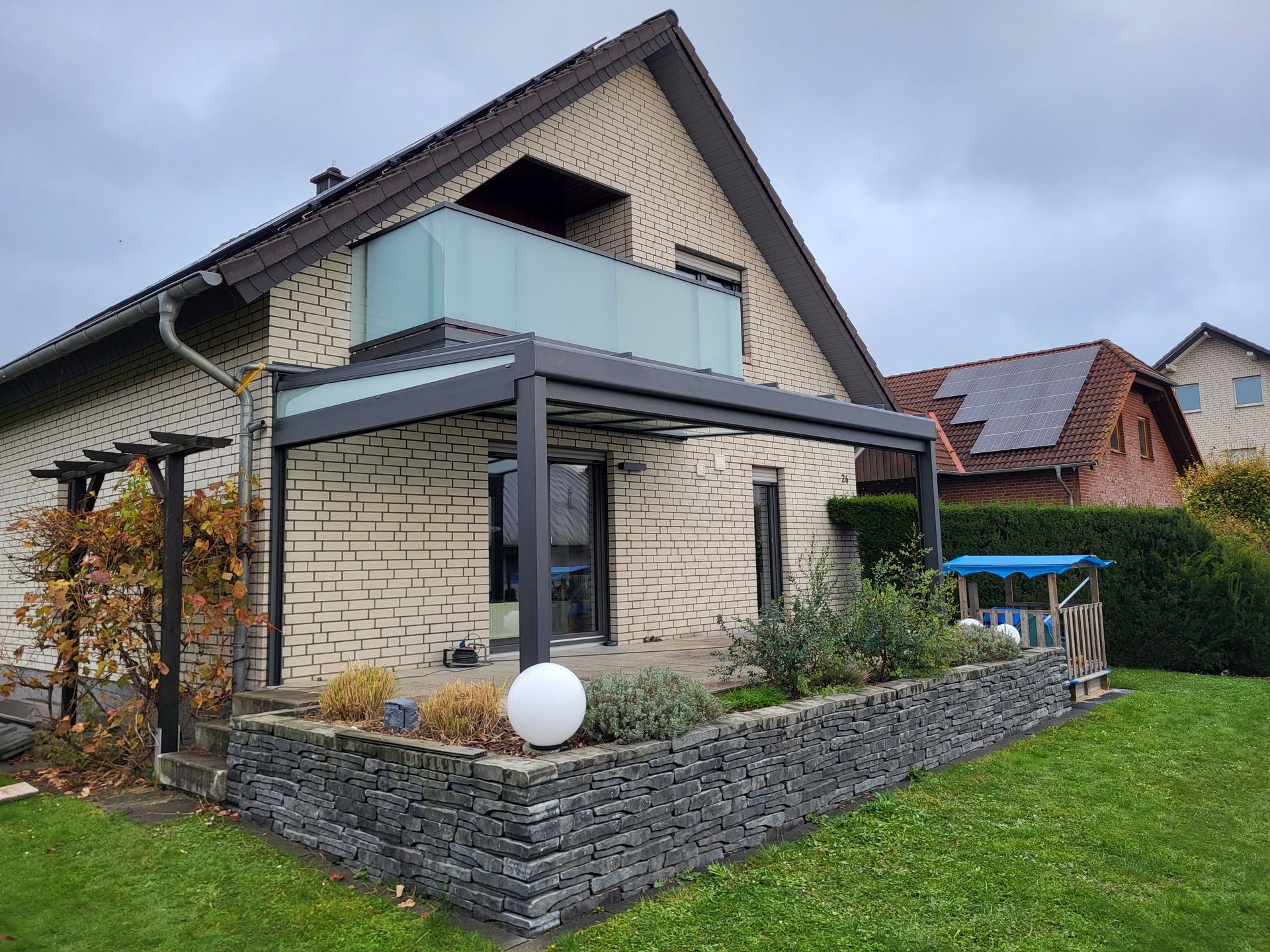 Modernes Haus mit Steinfassade, Balkon und überdachter Terrasse in einem angelegten Garten unter bewölktem Himmel