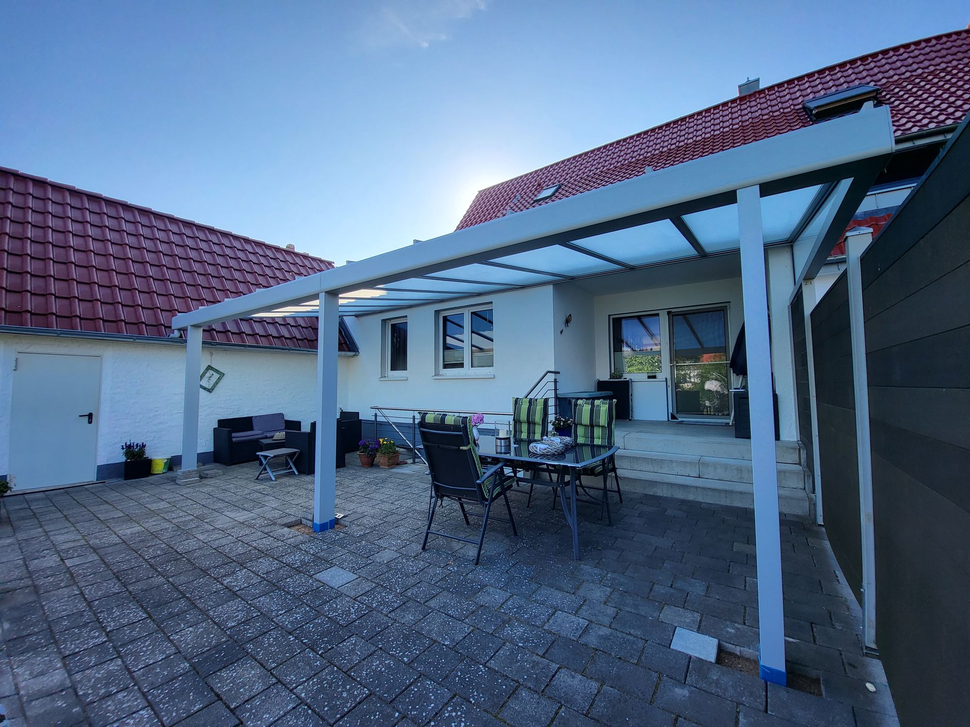 Terrasse mit Gartentisch und Stühlen neben einem weißen Haus mit rotem Ziegeldach und Glasmarkise