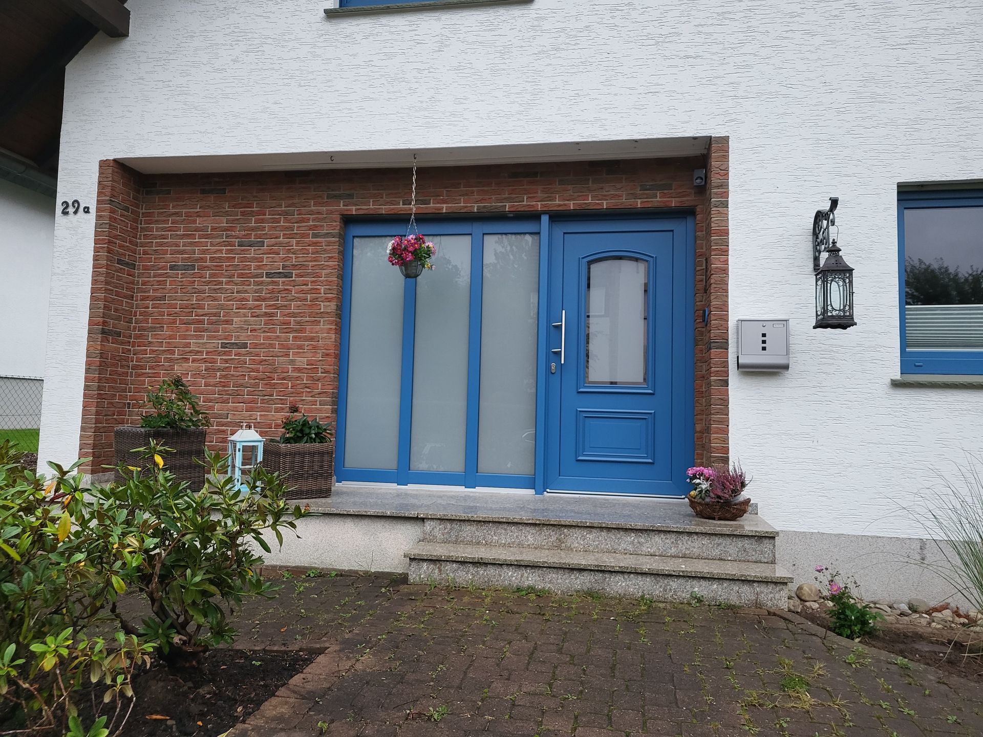 Blaue Eingangstür und Fenster an einem weißen Backsteinhaus mit kleiner Veranda und hängendem Blumenkorb.