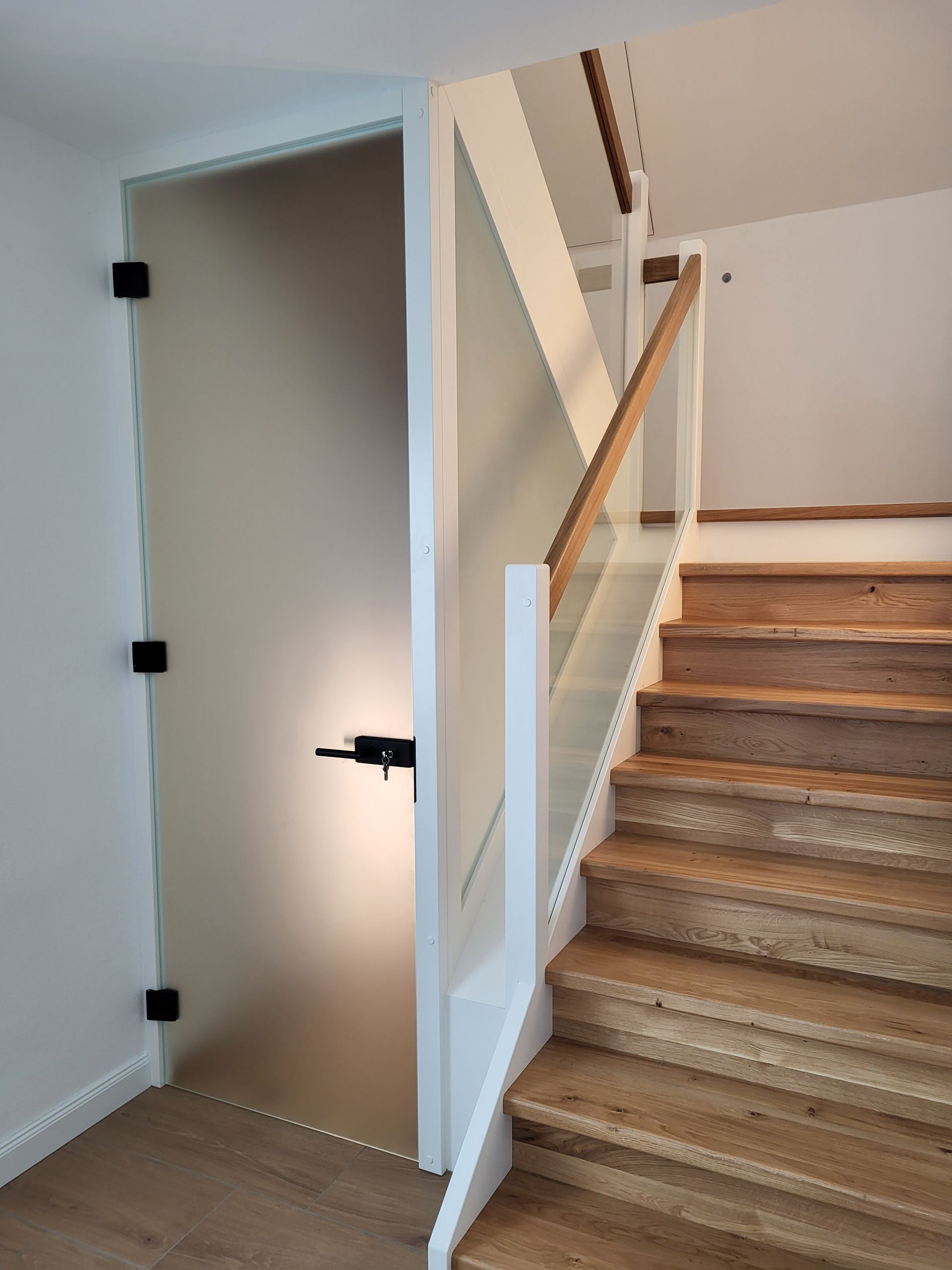 Treppe neben einer offenen Milchglastür mit schwarzen Scharnieren und einer Wandleuchte