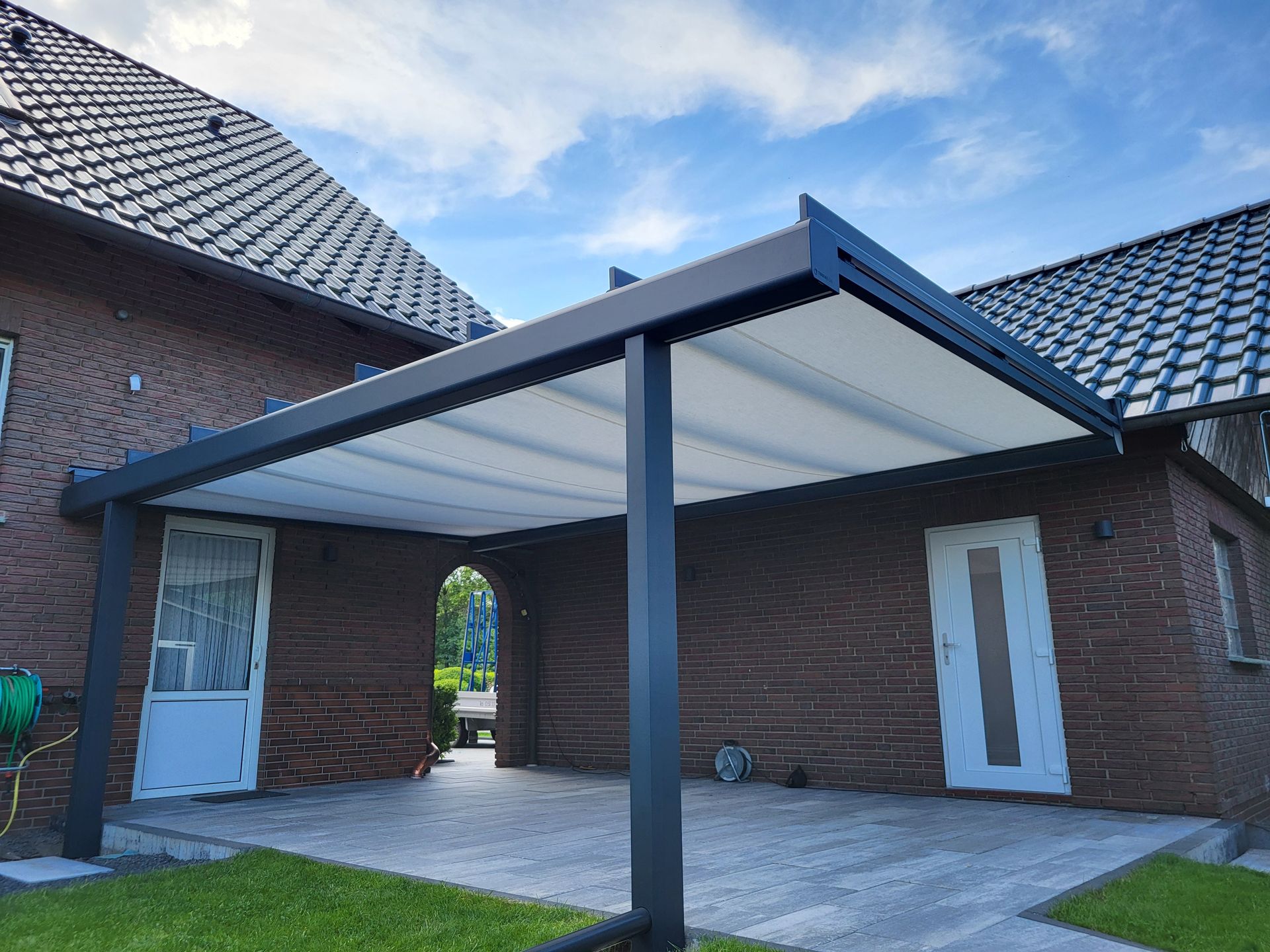 Überdachte Terrasse mit einer dunklen Metallpergola zwischen zwei Backsteinhäusern unter bewölktem Himmel.
