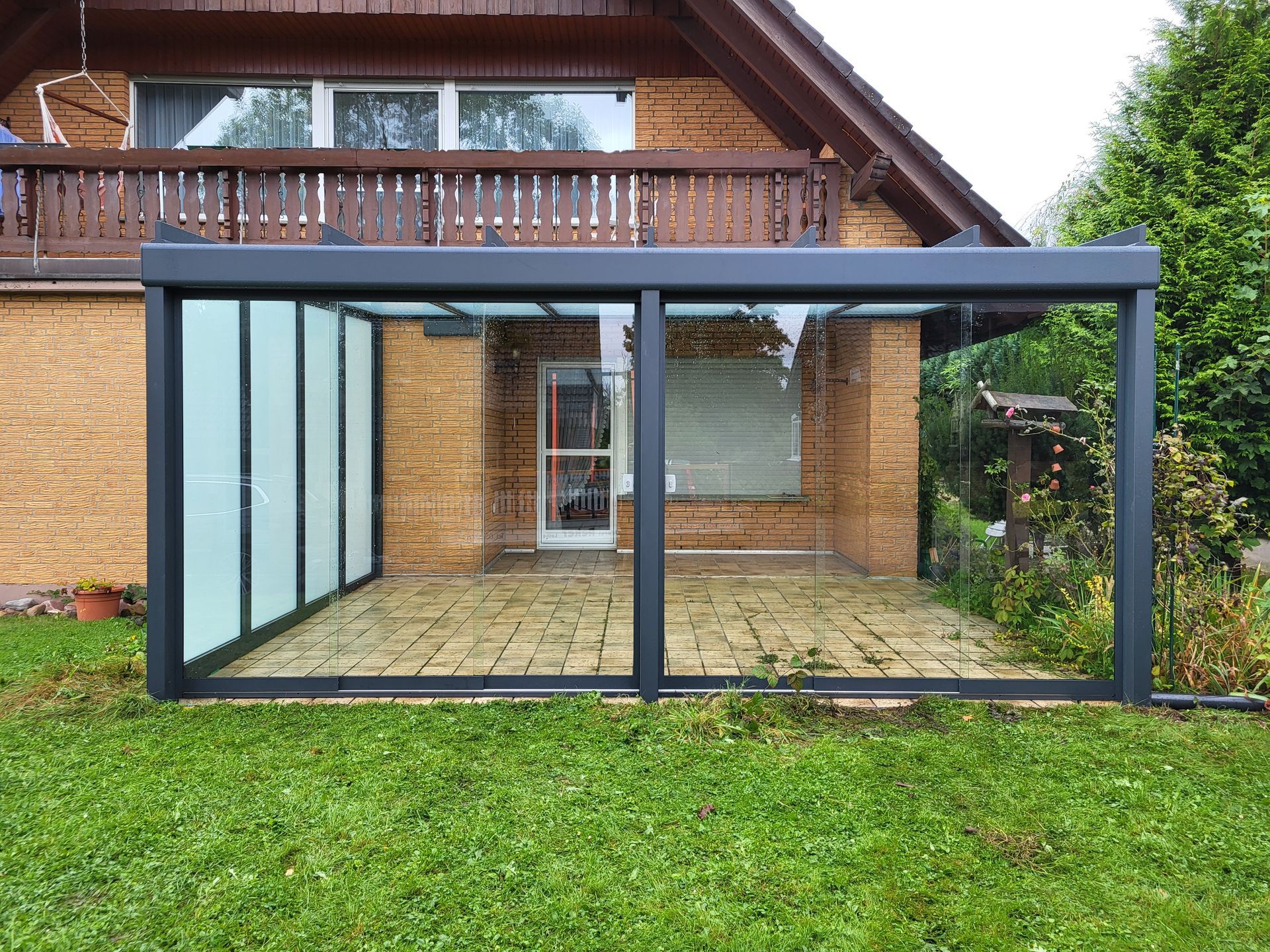 Eine verglaste Terrassenüberdachung mit dunklem Metallrahmen und Rasenfläche ist an ein Backsteinhaus angebaut.