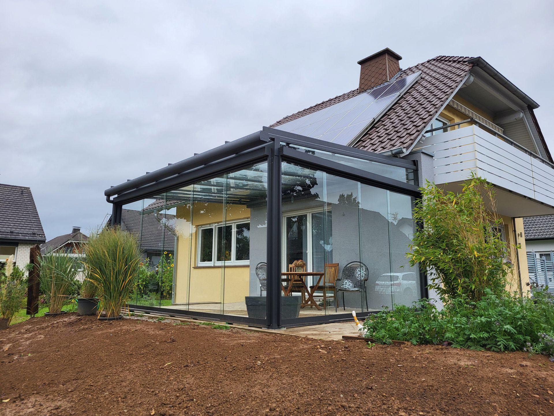 Modernes Haus mit verglaster Terrassenüberdachung und braunem Mulchgarten unter bewölktem Himmel