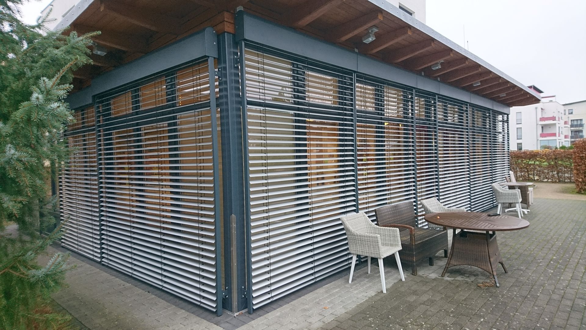 Moderne, mit Latten bespannte Terrasse neben einem Gebäude, mit Tisch, Stühlen und einem Wellblechdach.