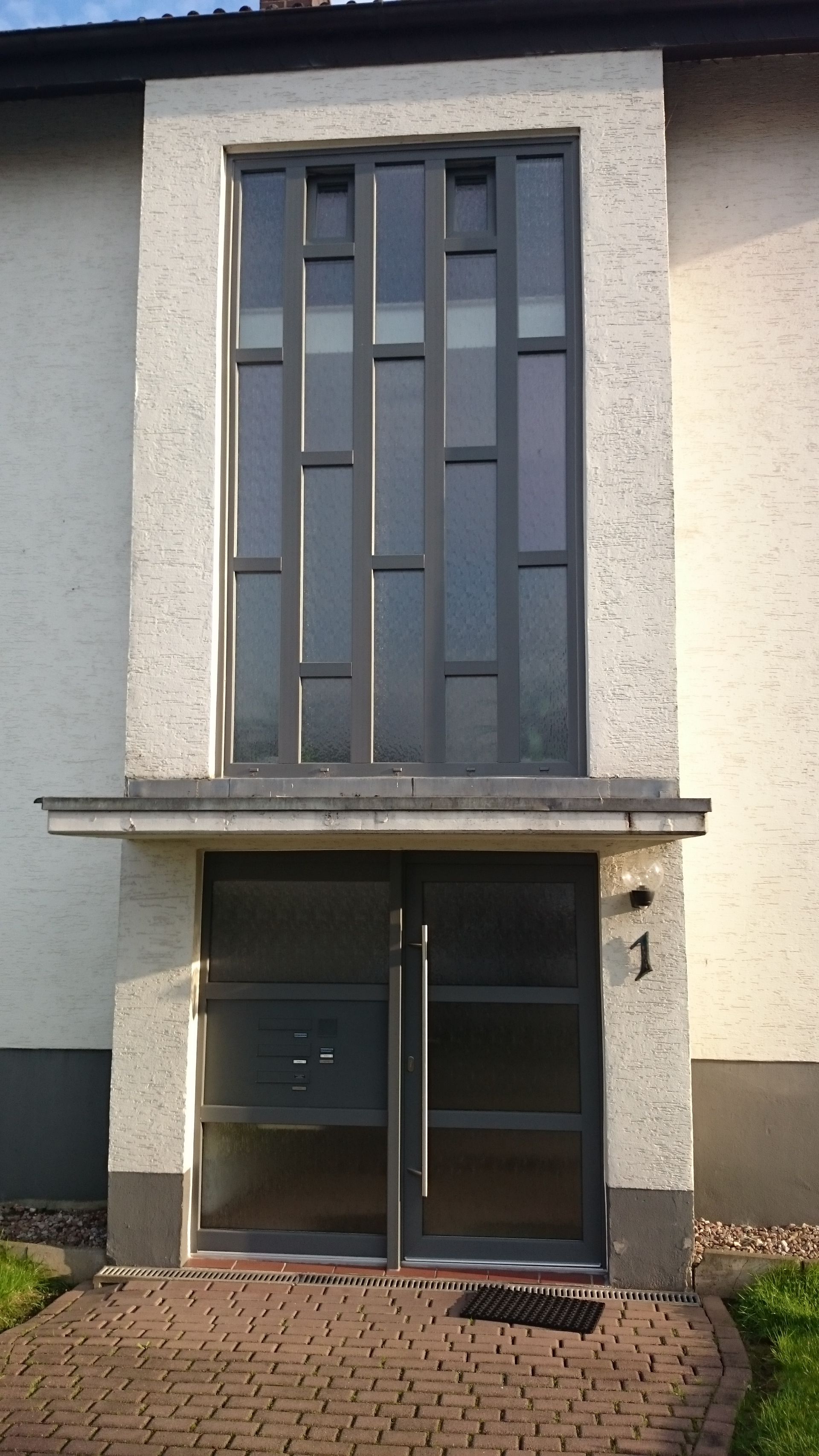 Vordereingang eines Hauses mit einem hohen, dunklen Fenster, doppelten Glastüren und einer kleinen überdachten Veranda.