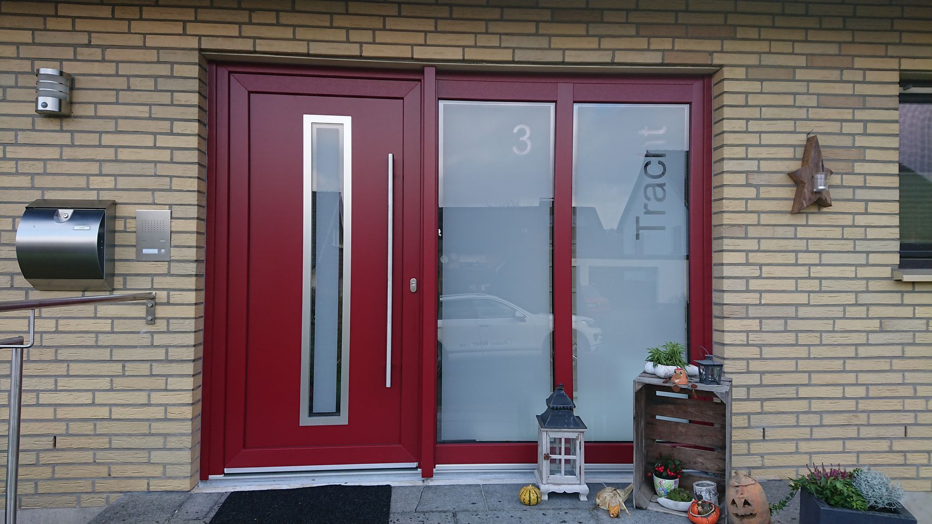 Rote Haustür mit schmaler Glasscheibe neben einem mattierten Seitenfenster an einer Backsteinfassade.